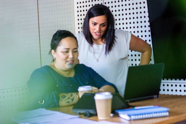 Two women collaborating at a desk with a laptop, takeaway coffee cups, and notebooks. One is seated, the other leaning over to look at the screen, both focused on their work.